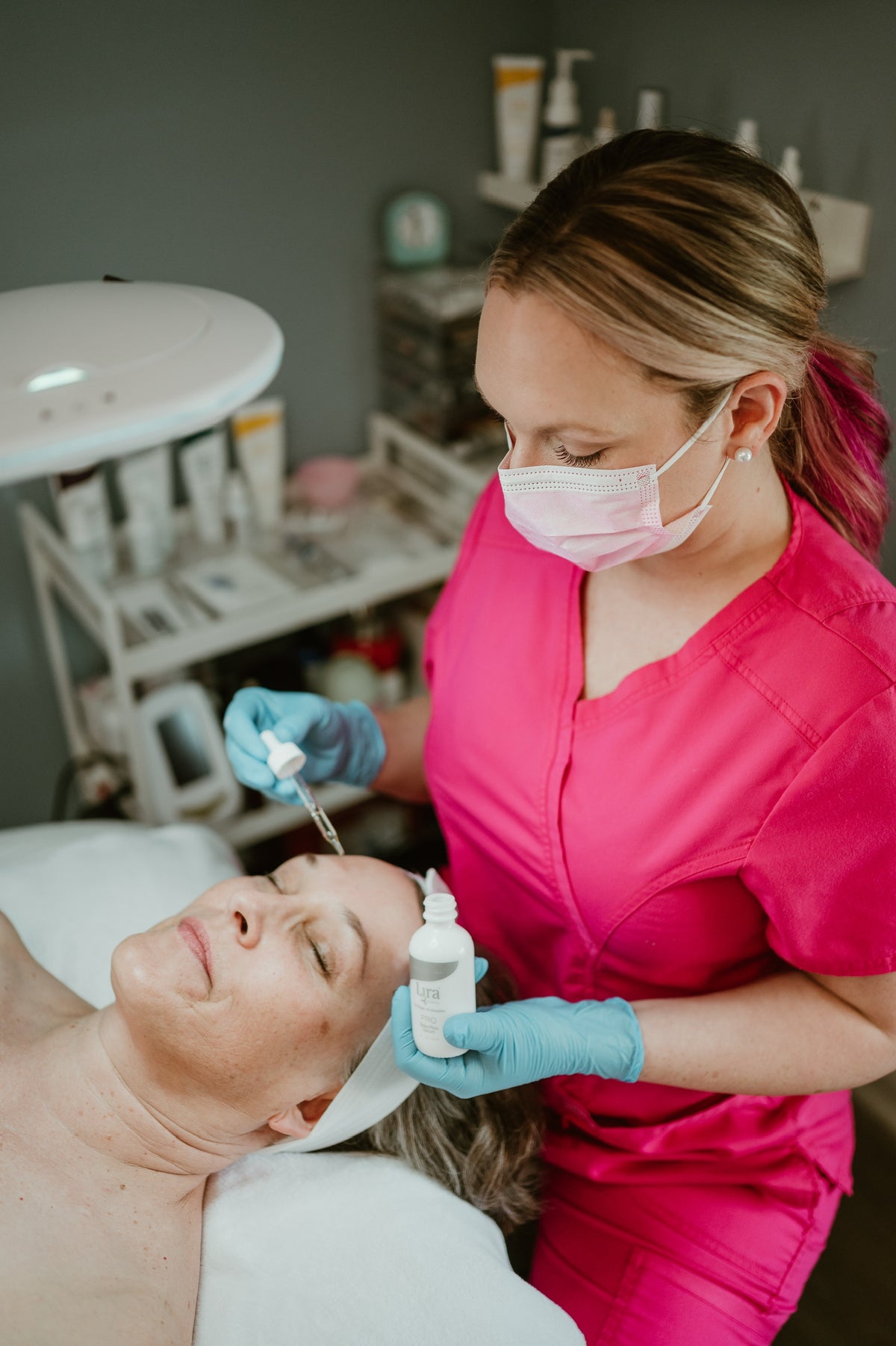 A beauty therapist in a bright pink scrub top and face mask applies a treatment to a client's forehead in a spa or clinic setting.