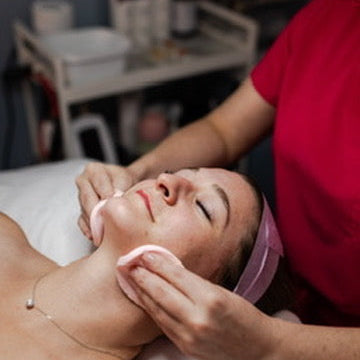 A woman receives a facial cleansing treatment with a spa worker applying a pink cleanser pad to her face in a treatment room.