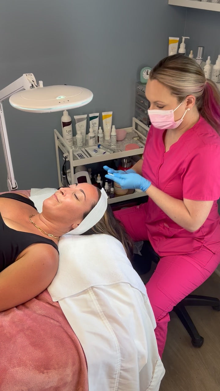 A woman in a pink scrub top and face mask attends a facial treatment while lying on a treatment bed under a bright lamp in a spa or beauty salon.