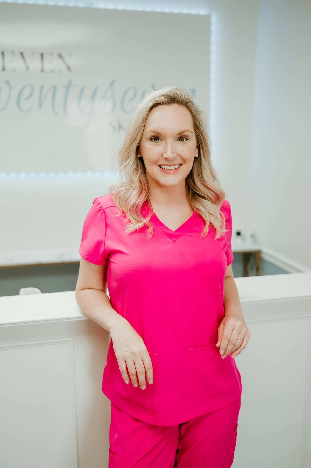 A smiling blonde woman wearing a bright pink scrub top and matching scrub pants stands indoors in a clean white clinical or spa setting.