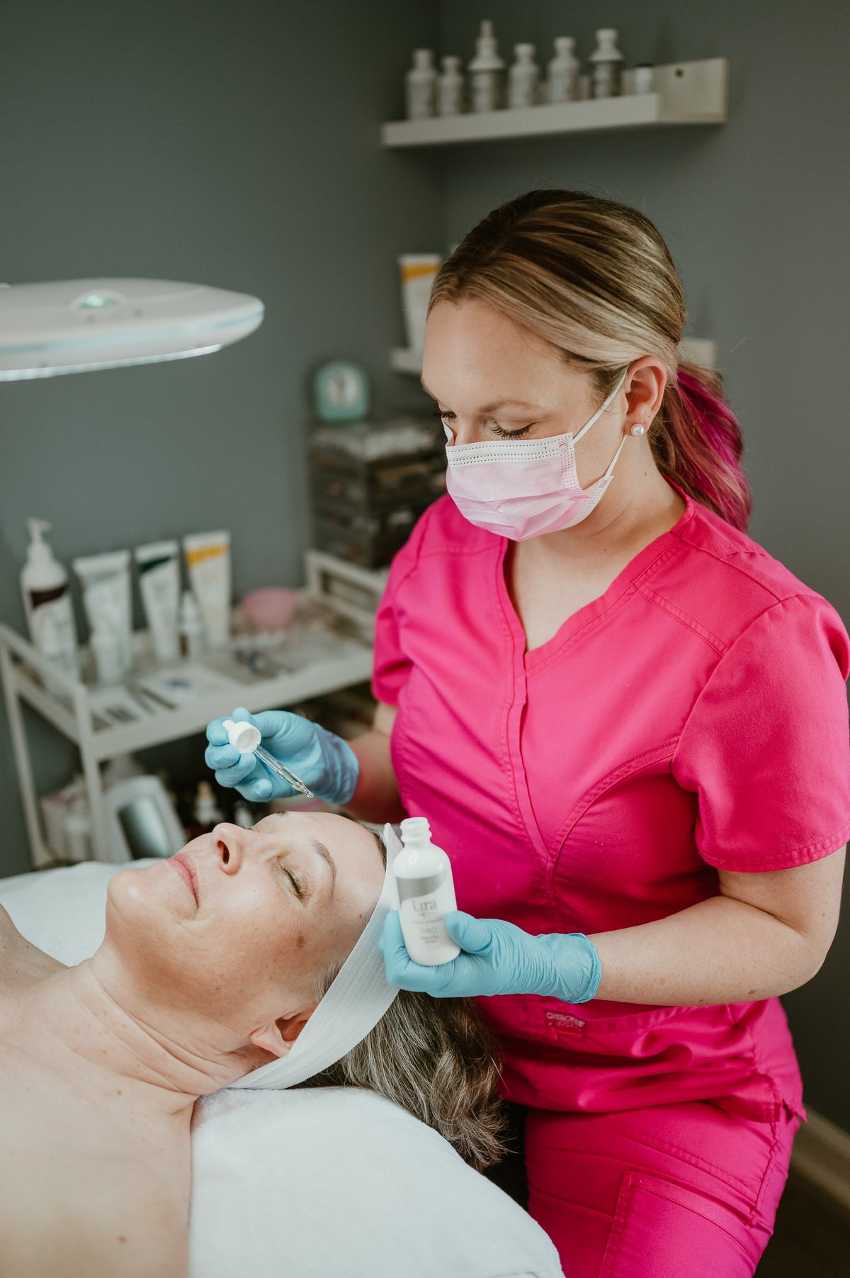 A beauty therapist in a bright pink scrub top and blue gloves applies a treatment to a client's forehead in a spa setting.