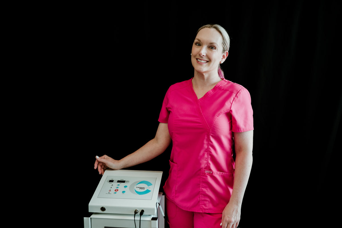 A woman wearing a bright pink medical scrub set stands next to a white medical machine against a solid black background.