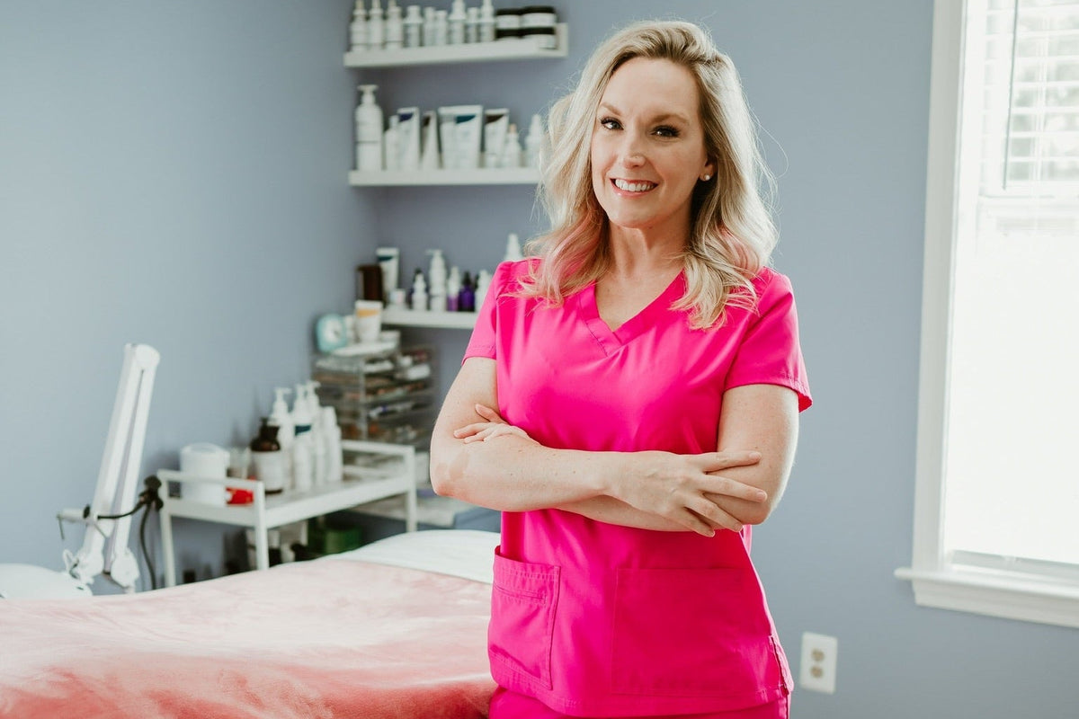 A woman wearing a bright pink medical scrub set stands with arms crossed in a brightly lit spa or treatment room with a bed and shelves of products in the background.