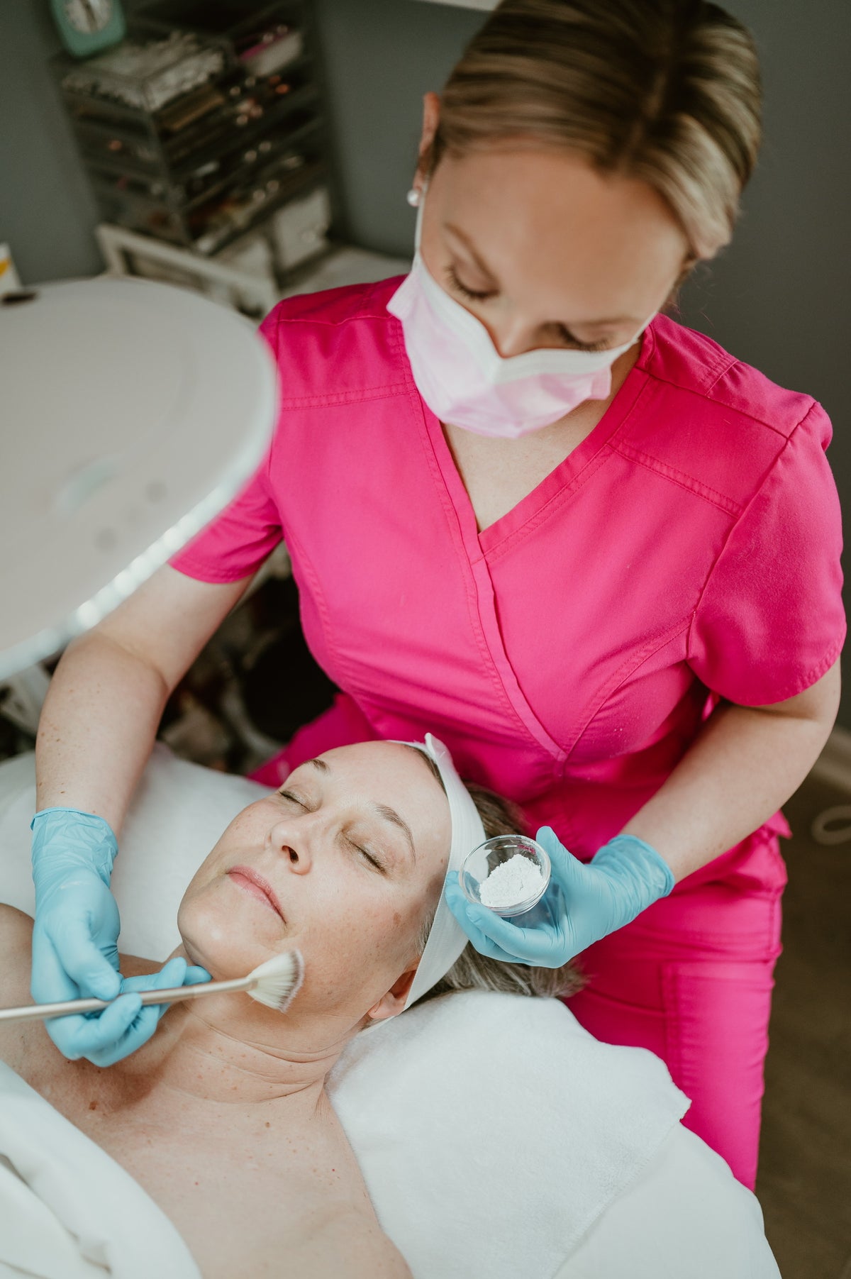 A beauty therapist in a bright pink scrub top and blue gloves applies a facial treatment to a client lying down on a treatment bed in a spa or clinic setting.