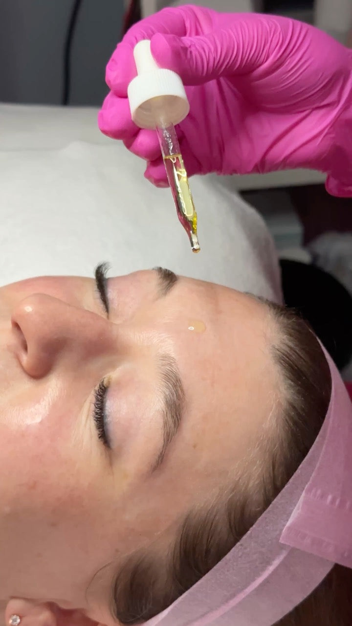 A close-up of a person lying down with their face covered in a pink headband, while a hand in a bright pink glove holds a glass dropper bottle above the forehead, suggesting a skincare or beauty treatment.