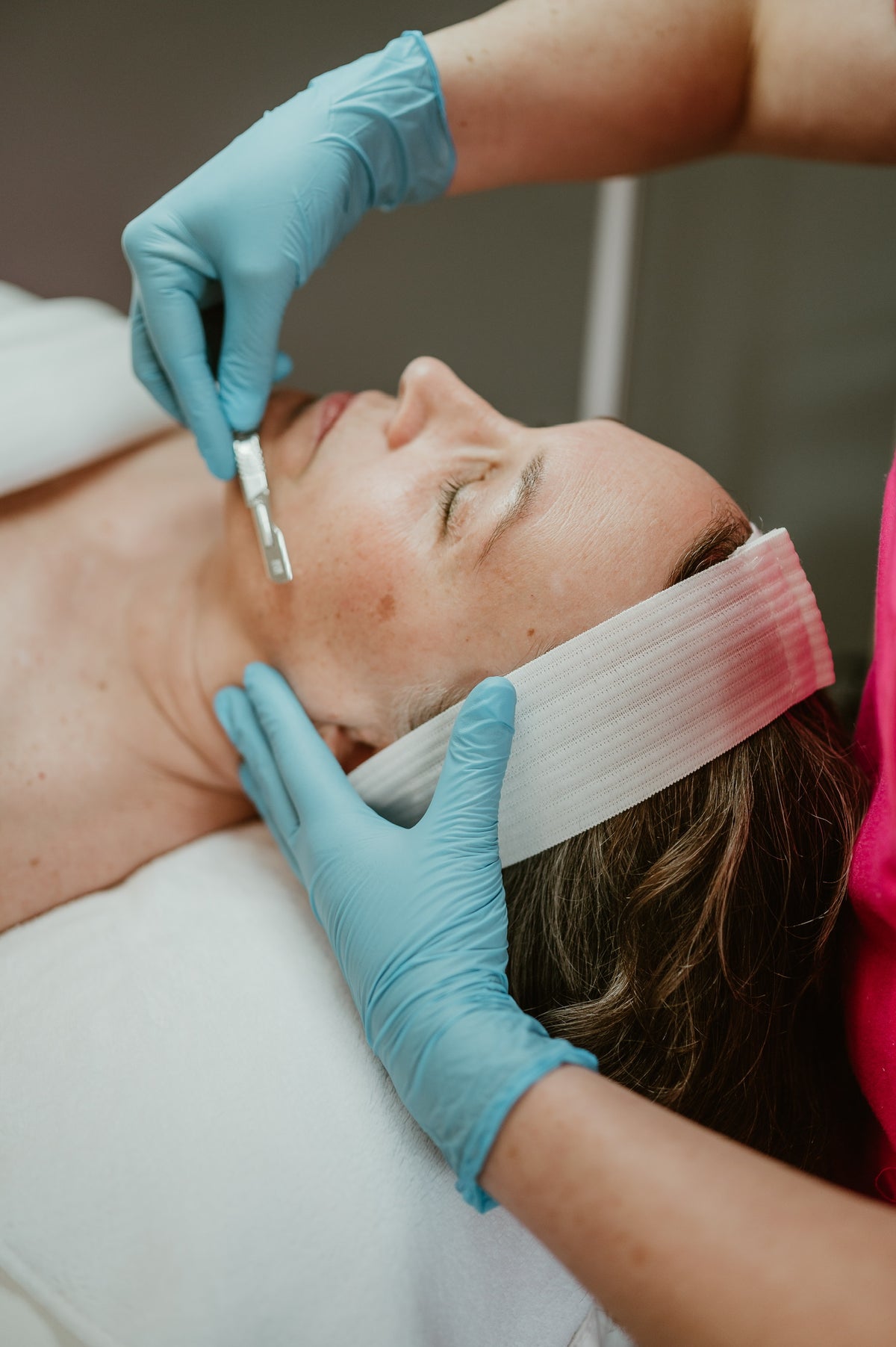 A close-up of a person lying down while another person in blue gloves applies a facial scrub or mask to the face. A white headband with pink tint is wrapped around the head.