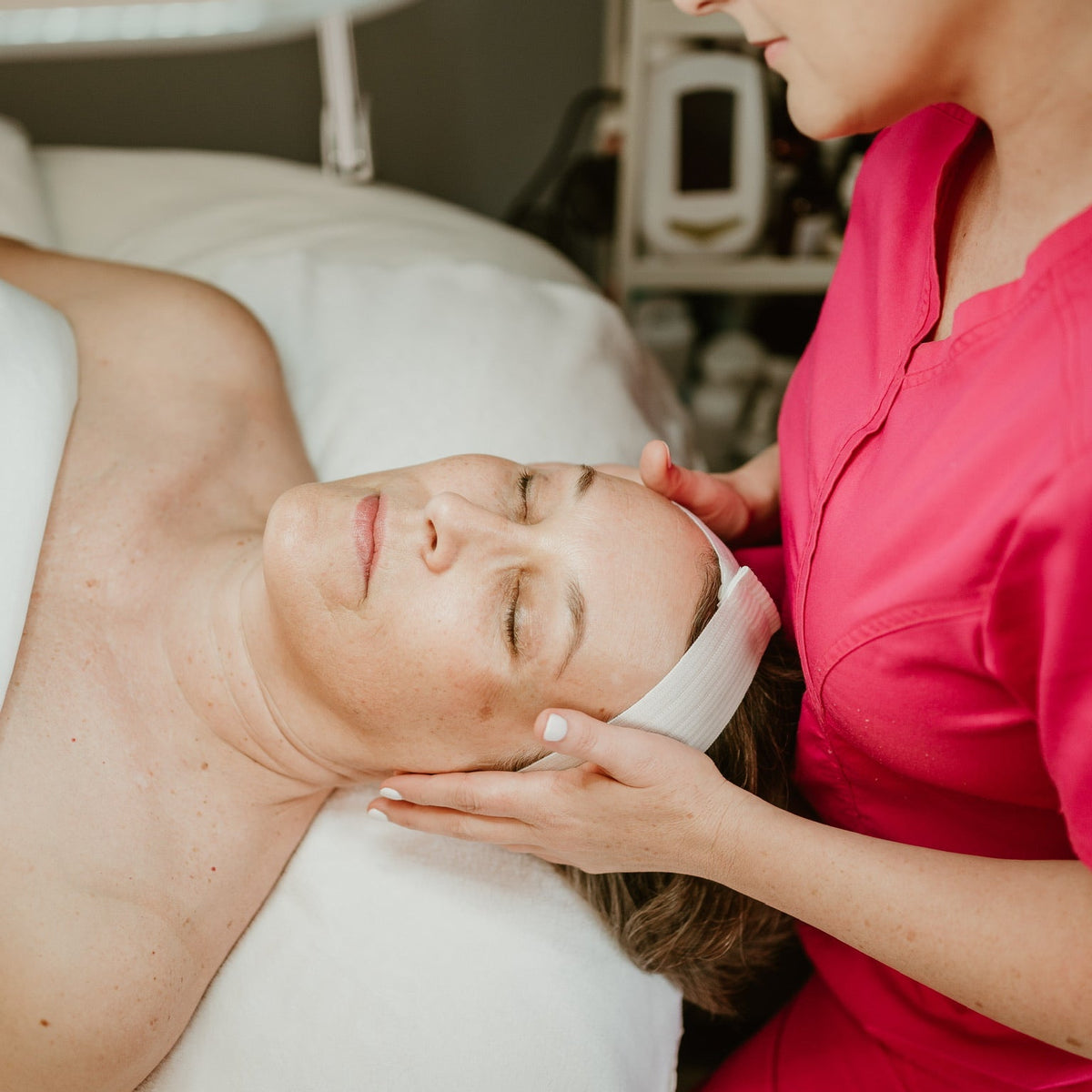 A woman lies on a treatment bed with her eyes closed while a provider in a bright pink uniform gently massages her forehead. A white headband is wrapped around her head, and spa equipment is visible in the softly blurred background.
