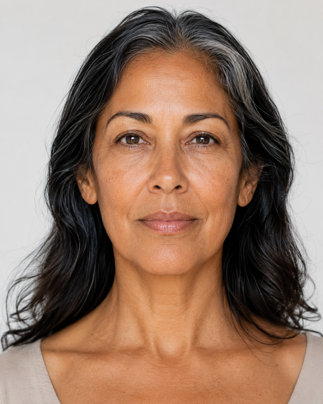 A close-up portrait of a woman with long, wavy black and gray hair, wearing a light beige top against a plain light gray background.