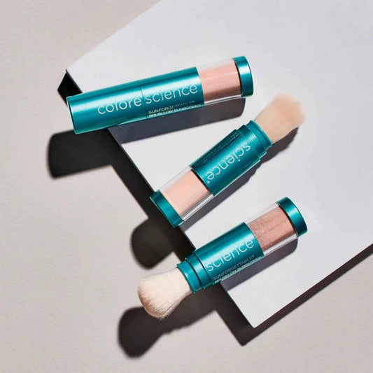 Three Colore Science Sunforgettable Total Protection Brush-On Shield SPF in teal tubes, with brush tips, arranged on a white geometric surface against a light gray background.