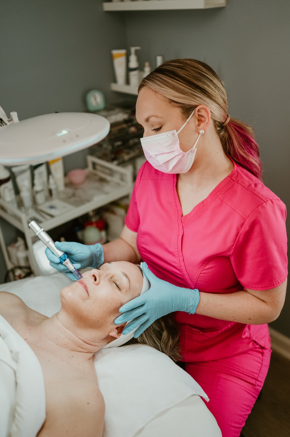 A beauty therapist in a bright pink scrub top and blue gloves applies a cosmetic treatment to a client's face while the client lies on a treatment bed in a spa or clinic setting.