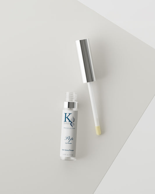 A clear cosmetic bottle with a silver cap and applicator wand, shown on a clean light gray background. The bottle label has a blue 'K' logo and text, and the applicator tip contains a pale yellow cream.