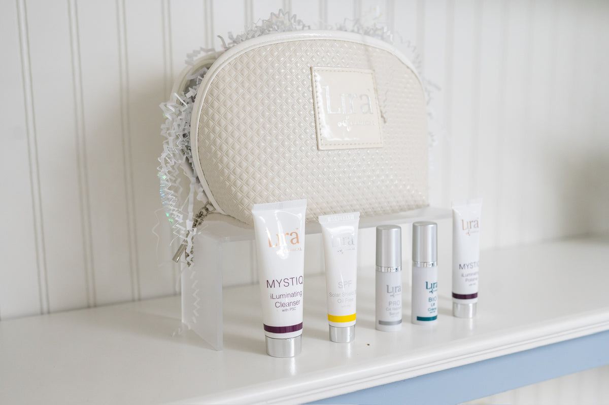 A white textured cosmetic bag with fringe trim and five skincare tubes from the brand L'ya, displayed on clear acrylic risers on a white shelf against a white paneled wall.