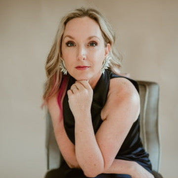 A woman with blonde hair and pink streaks sits in a chair wearing a black sleeveless top and large silver earrings against a neutral beige background.