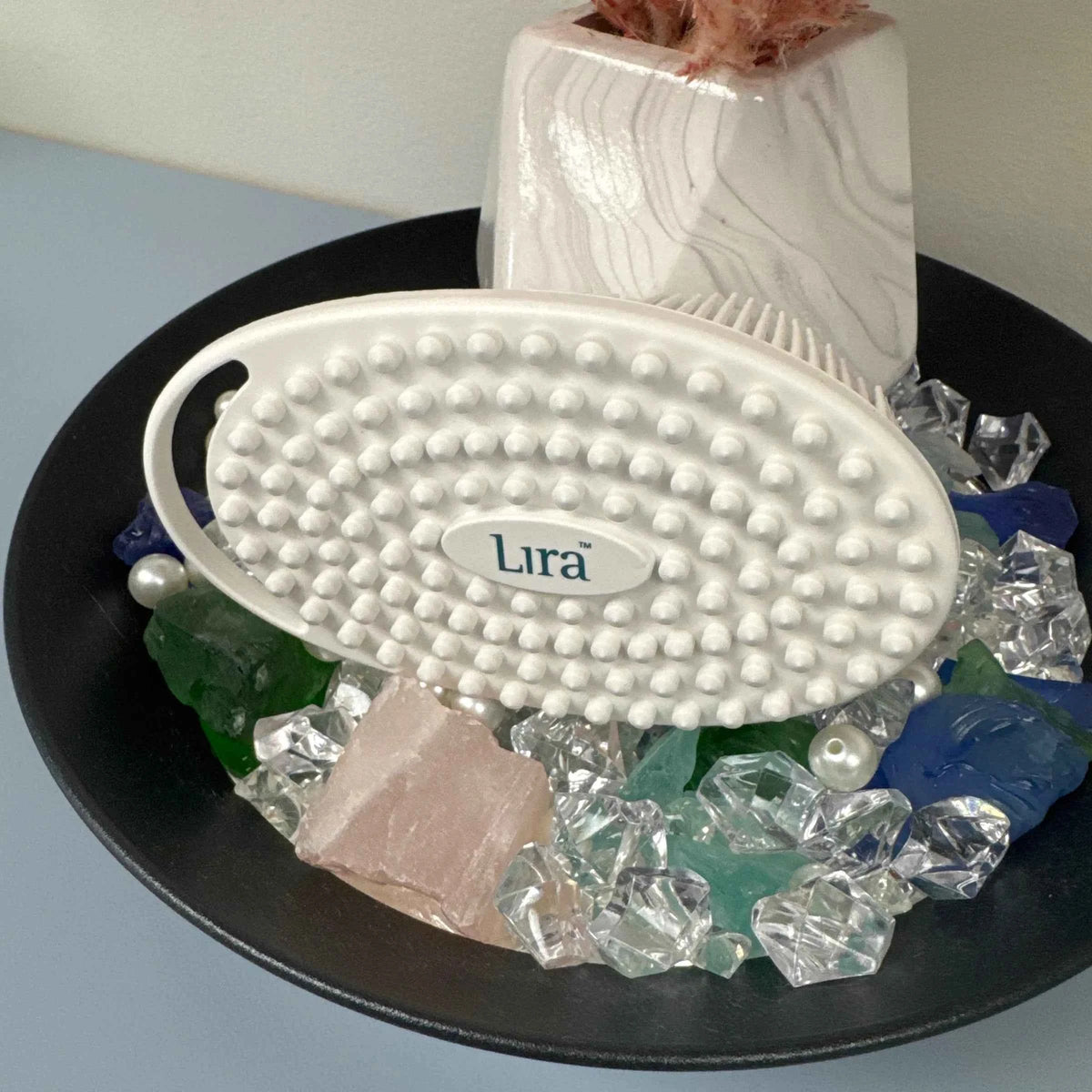 An oval white hairbrush with nubs and a visible 'Lira' logo, displayed on a black tray with decorative crystals and a white vase in the background.