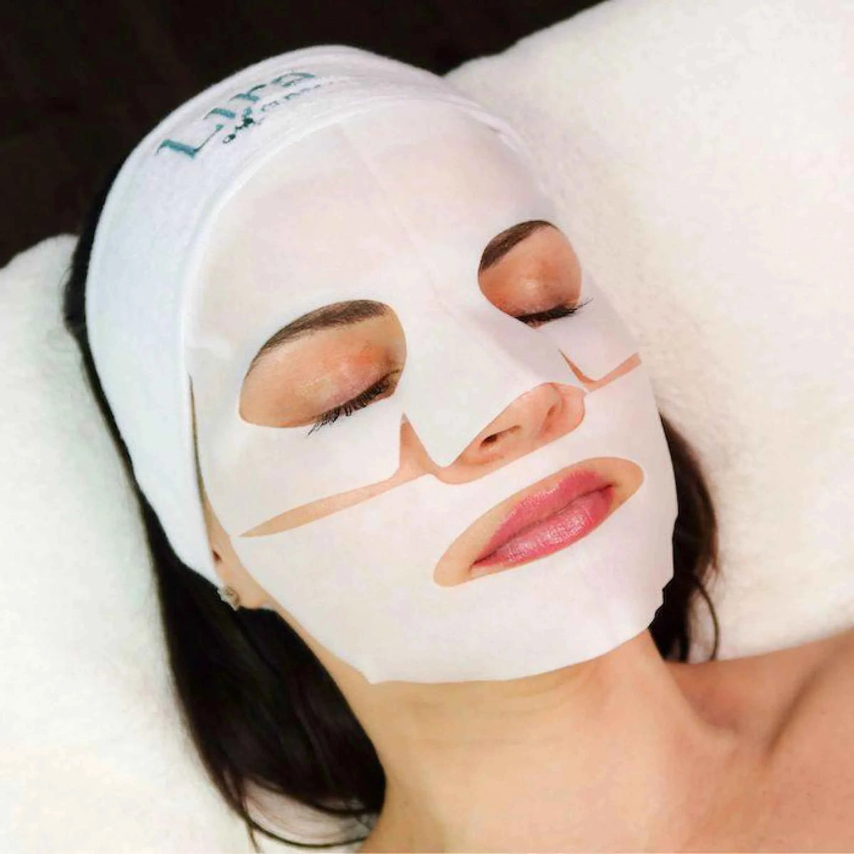 Woman reclining with eyes closed while wearing a white facial sheet mask and Lira brand head band, against a white cushion and dark background.