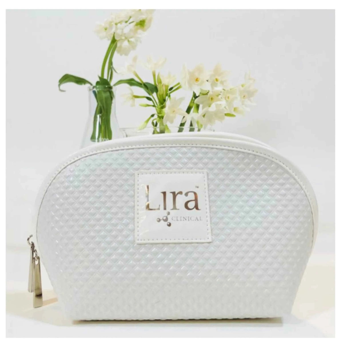 A white textured cosmetic pouch with a white leather patch reading 'Lira Clinical' on the front, shown with a gold zipper pull and a small vase of white flowers behind it on a clean white background.