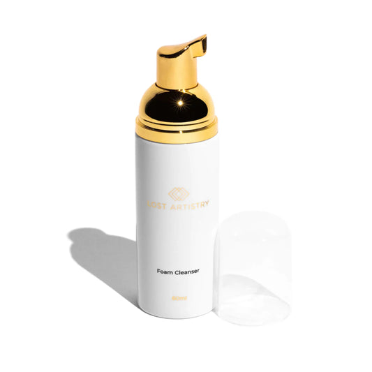 A white cylindrical foam cleanser bottle with a shiny gold pump dispenser and gold logo text, shown on a clean white background with its clear cap placed beside it.