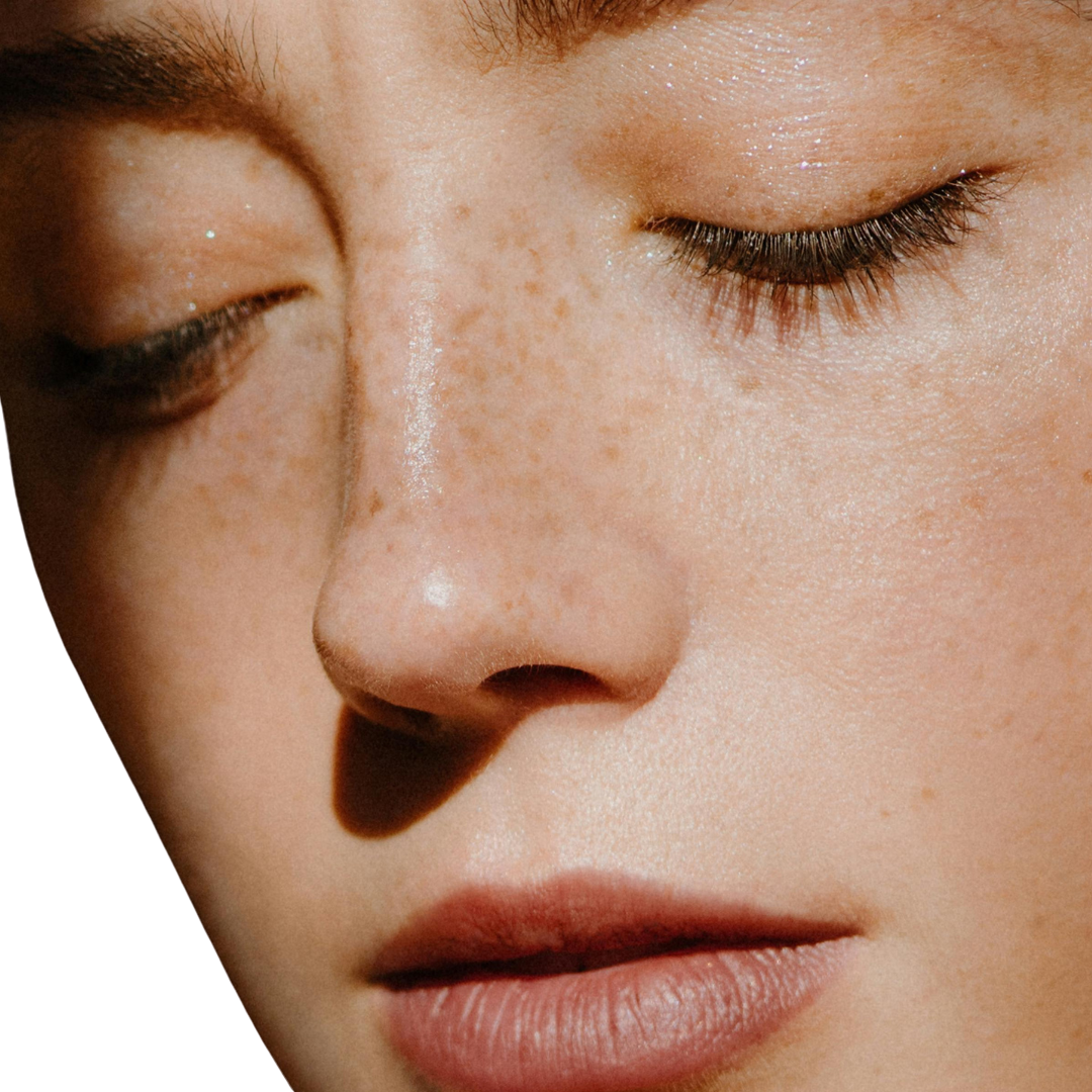 Close-up of a person's face with closed eyes, showing natural skin texture, freckles, and soft shadowing against a white background.