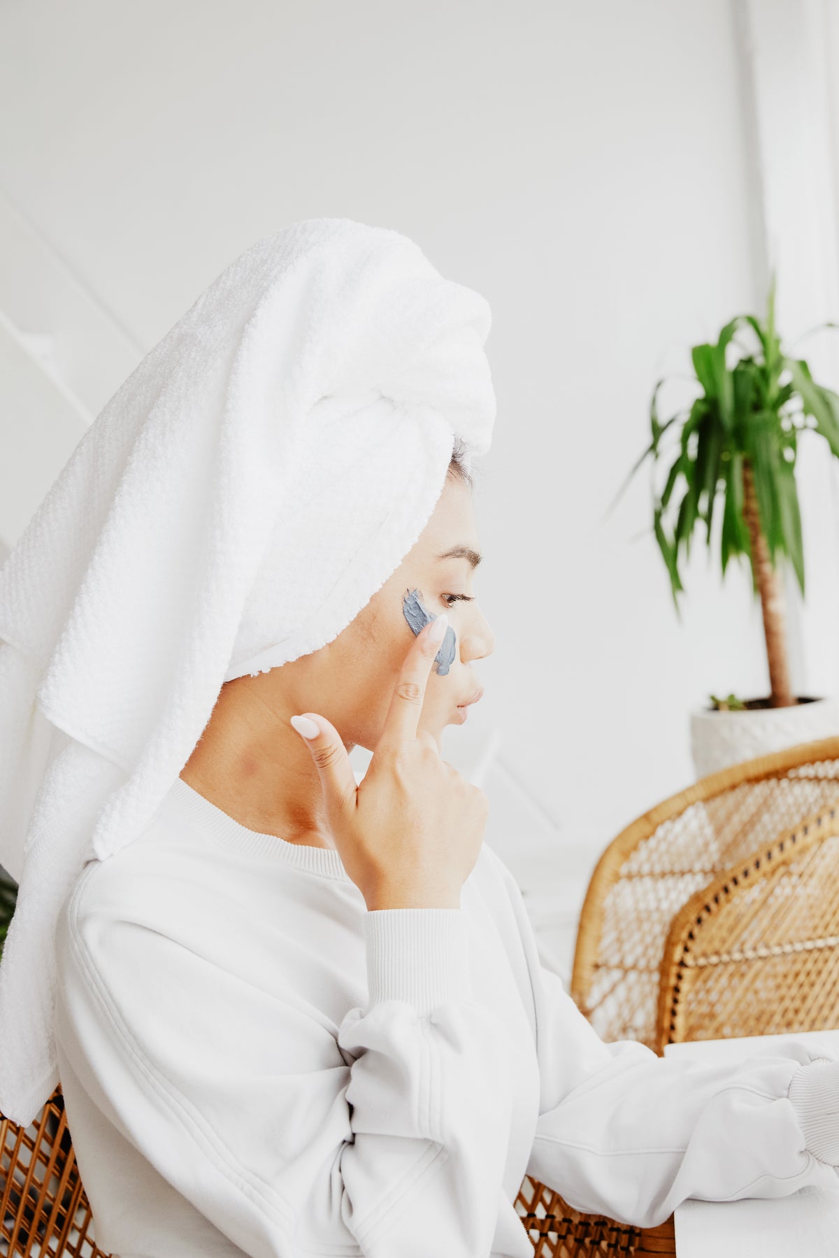 Woman with a white towel wrapped around her hair applying a facial skincare product to her cheek while seated in a bright, spa-like room.