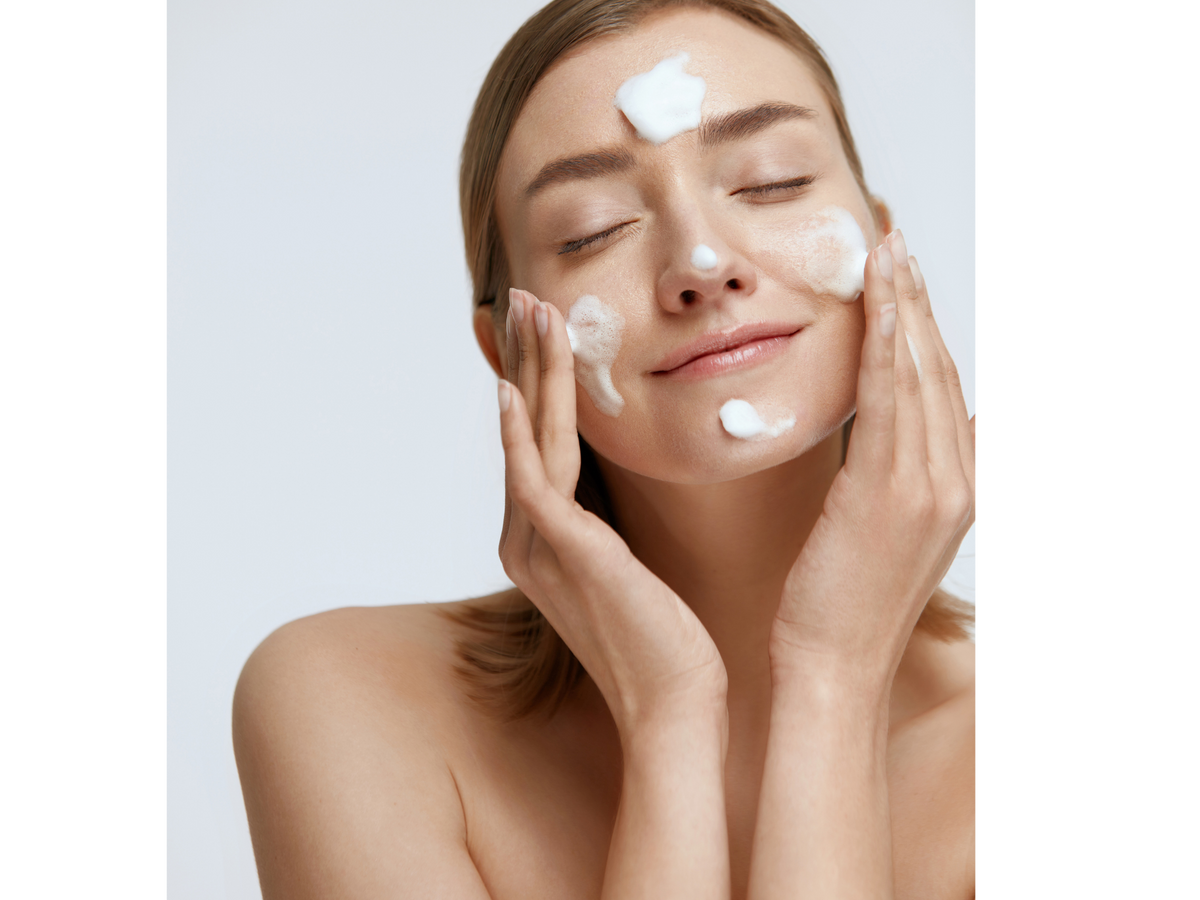 A woman with closed eyes applying or washing white face cream onto her face and hands, shown in a close-up portrait against a light gray background.