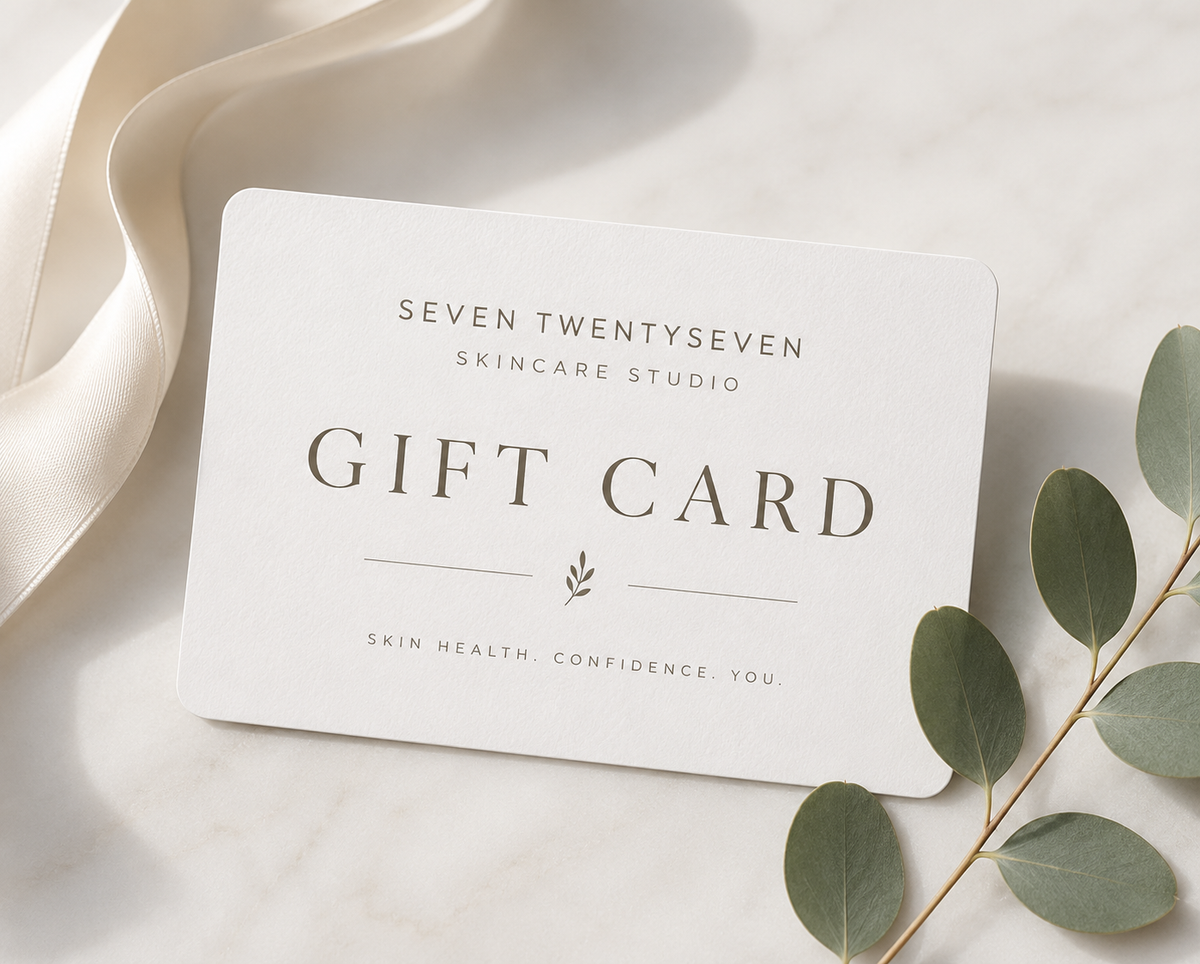 A white gift card with elegant gray text reading 'SEVEN TWENTYSEVEN SKINCARE STUDIO GIFT CARD' and 'SKIN HEALTH CONFIDENCE YOU,' styled on a light marble surface with a cream ribbon and eucalyptus leaves.