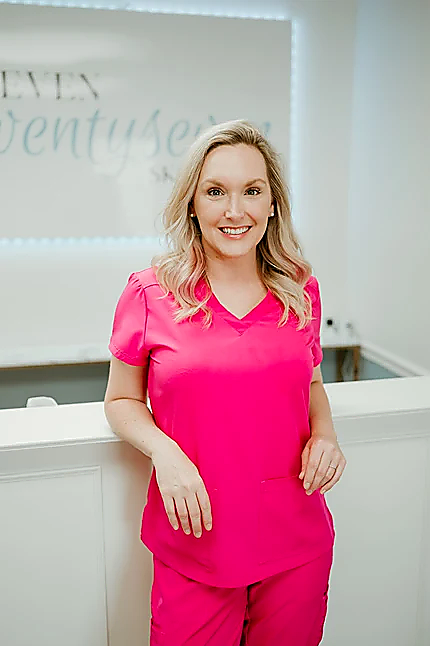 A smiling blonde woman wearing bright pink medical scrubs stands indoors against a white wall with a sign in the background.