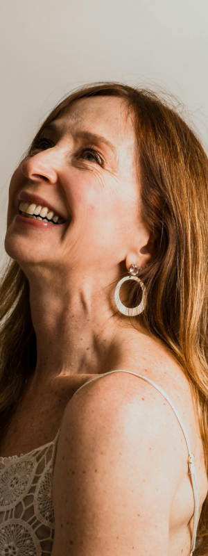 A close-up portrait of a smiling woman with long light brown hair wearing a large silver-toned hoop earring with a textured, stone-like border.