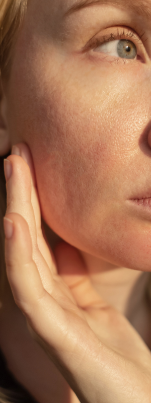 Close-up of a woman's face and hand under warm sunlight, showing natural skin texture and a soft-focus background.
