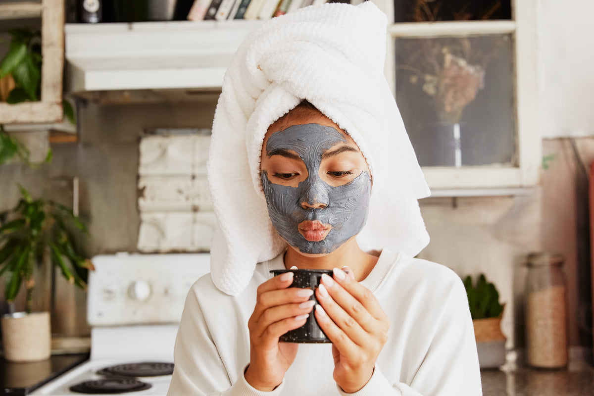 Woman wearing a white towel wrap and gray facial mask holding a mug during an at-home skincare routine.